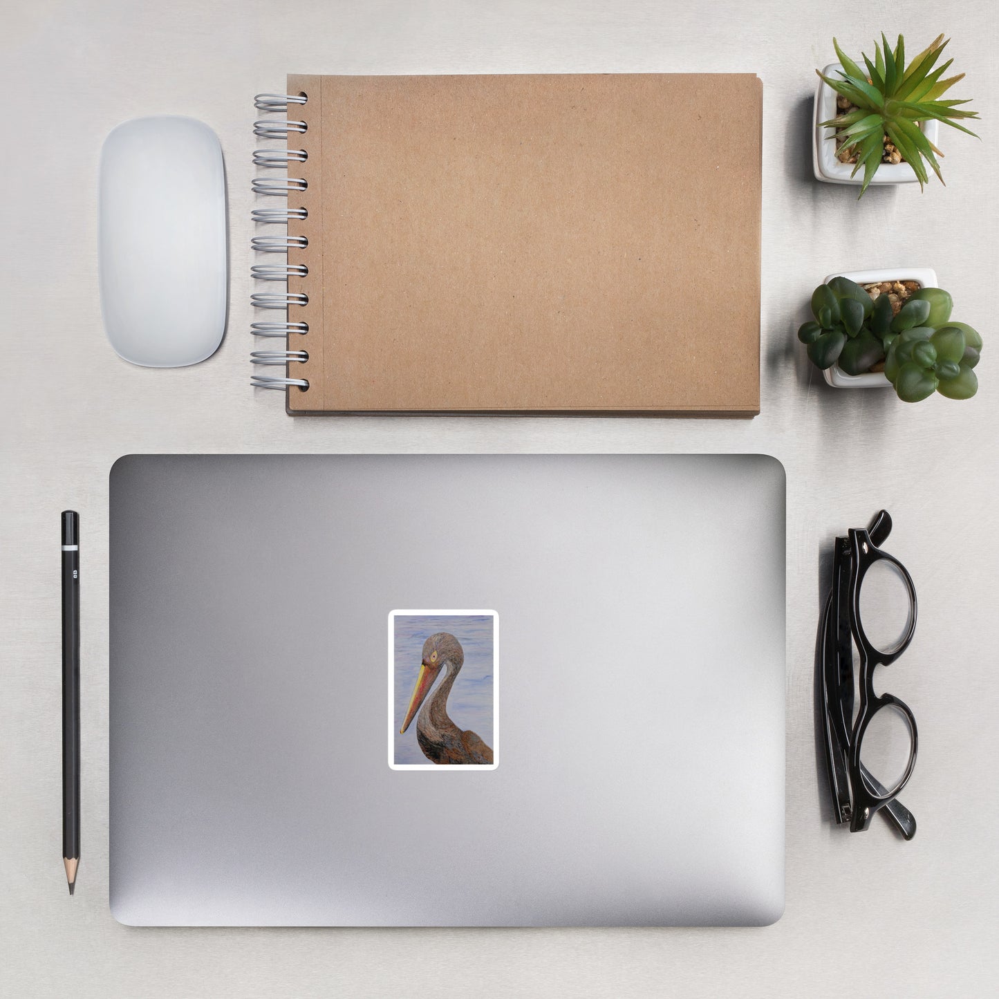 Laptop with a Louisiana pelican sticker, notebook, mouse, pencil, glasses, and plants on a light surface